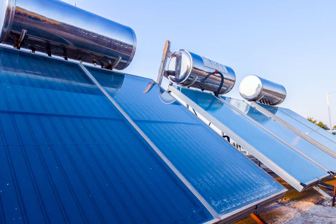 A solar water heater system is seen atop the slanted roof of a house complete with three large cylindrical water tanks and three expansive sections of blue tinted solar panels shown against a pale blue sky in the background, solar water heater, solar powered water heater, water heater, boiler, solar powered, solar, solar panels, roof, house, pale blue sky, blue sky, sky, pale blue, slanted roof, plumbing, electrical, hvac