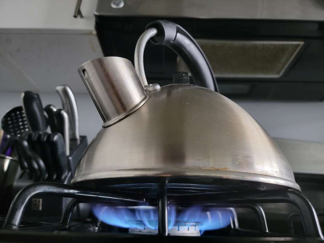 A stainless steel teapot sits atop one of the burners of a gas powered stove with a blue flame underneath