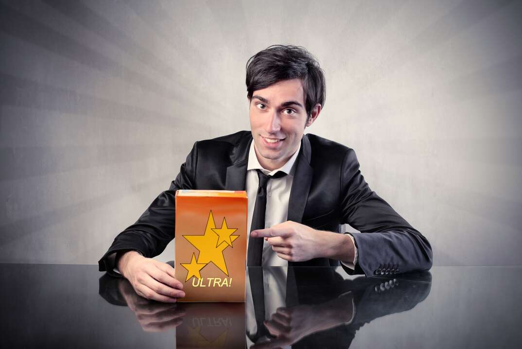 A shady looking man in a black suit and white shirt appears ostensibly in a TV advertisement pointing to a chintzy looking product in a box labeled with the brand name Ultra on it along with a gold star, TV, TV screen, TV ad, TV advertisement, TV commercial, commercial, ad, advertisement, advertising, huckster, sleazy salesman, salesman, shady, shady looking, black suit, black tie, white shirt, product
