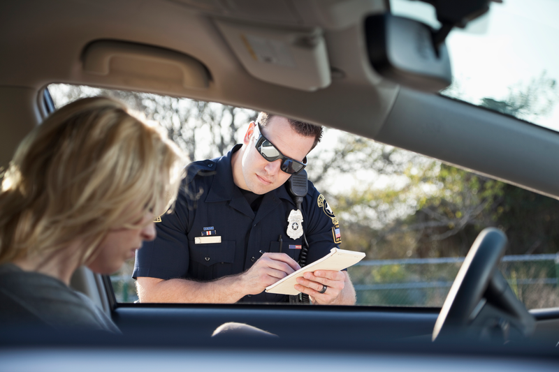 A uniformed police office wearing sunglasses writes a traffic citation to a blonde woman sitting behind the wheel of a stopped vehicle, uniformed police officer, police officer, cop, sunglasses, woman, blonde woman, blonde, stopped car, stopped vehicle, vehicle, car, ticket, traffic ticket, traffic, citation, traffic citation