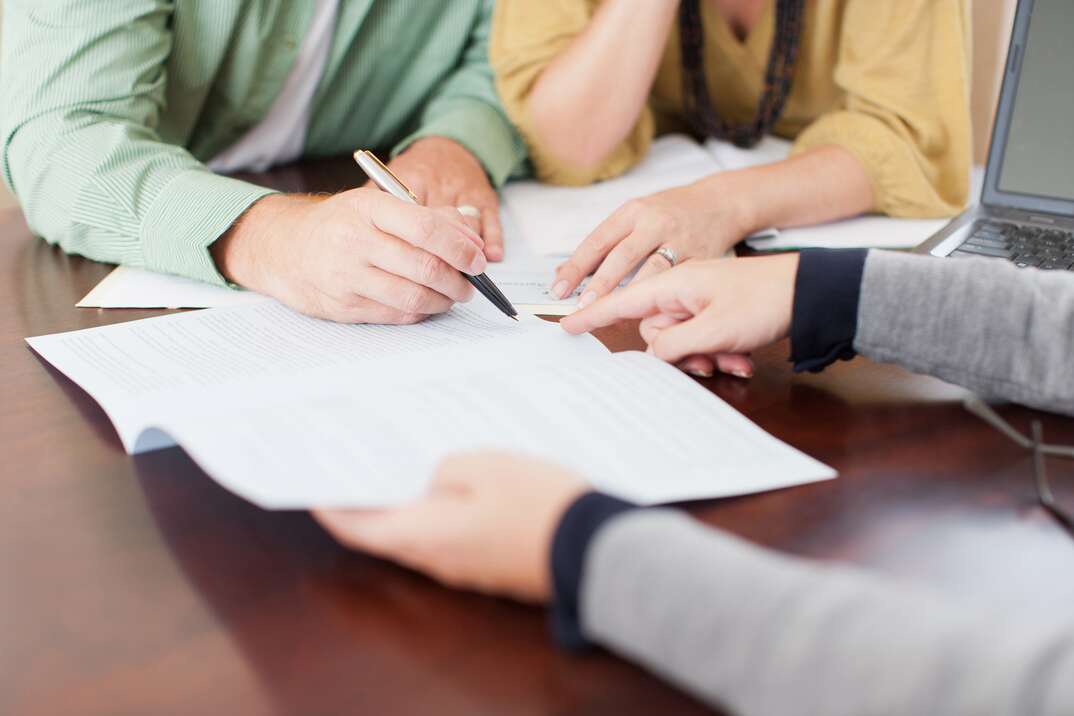 A married couple sits at a desk across from a lawyer as one of them signs a legal document, lawyer, attorney, law, legal, document, contract, papers, legal document, legal contract, legal papers, human hands, humans, married couple, married, hands, signing documents, signing contracts, signing papers, pen, ink pen, desk, legal office, law office, lawyer's office, attorney's office, man and woman, postnuptial agreement