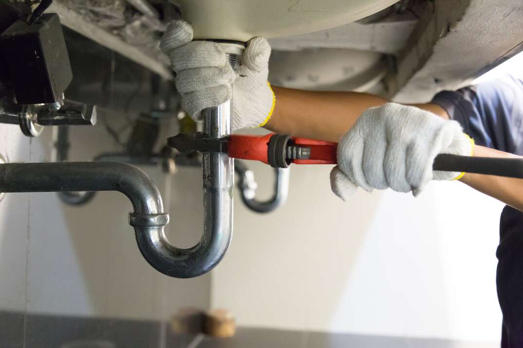 Person wearing rubber gloves uses an adjustable wrench on a drain pipe underneath a sink.