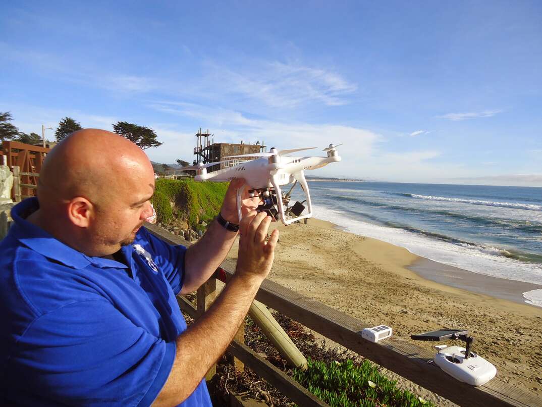 A bald man in a blue shirt stands near a California beach as waves from the Pacific Ocean roll in while he prepares to fly a drone on a clear day with blue skies, drone, drone pilot, drone operator, gadget, flying, remote control, beach, sand, beachfront, coastal, coast, West Coast, Pacific Ocean, California, waves, ocean, blue skies, skies, clear skies, clear sky, sky, blue sky, white clouds, clouds, bald, bald man, bald guy, blue shirt