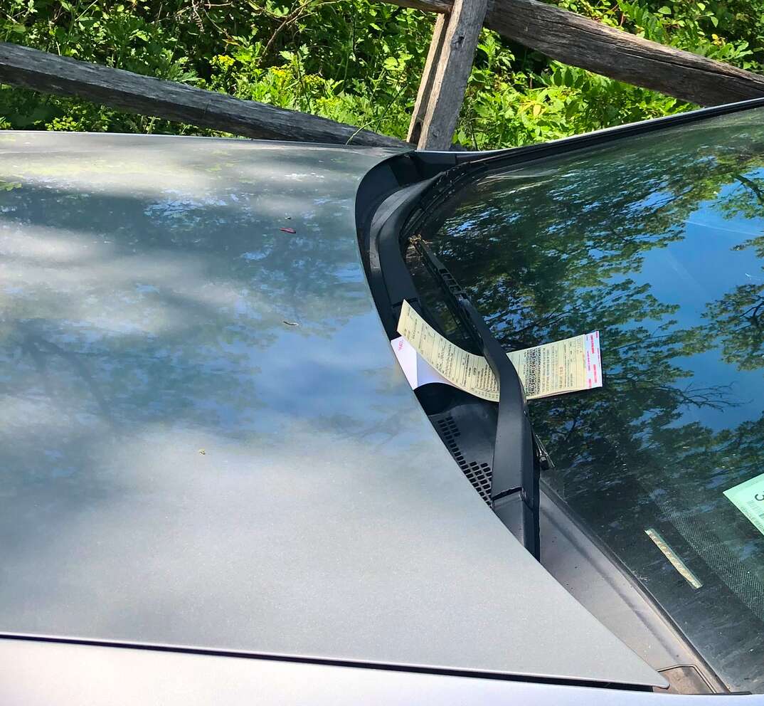 A parking ticket is secured under the windshield wiper of a car parked on the side of a road against a lush green natural background, parking ticket, ticket, parking, park, citation, cite, cited, illegal, legal, meter maid, meter, windshield, windshield wiper, wiper, hood, car hood, hood of car, greenery, parked, parking, park, ticket