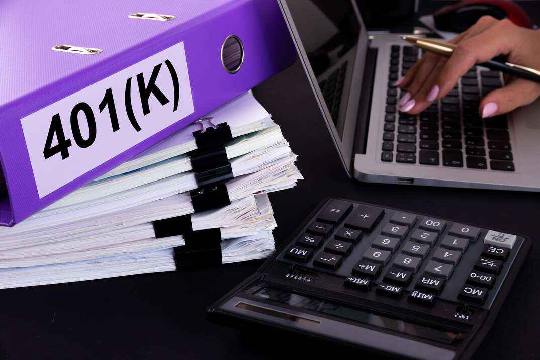 A woman's hand holds a pen as she rests her fingers on the keyboard of an open laptop computer that sits next to a black calculator and a stack of paper clipped documents with a purple binder marked 401K on top of them, 401K, 401(k), finance, financial documents, documents, papers, financial papers, financial, woman's hand, pen, calculator, desk, paper, binder, ring binder, purple