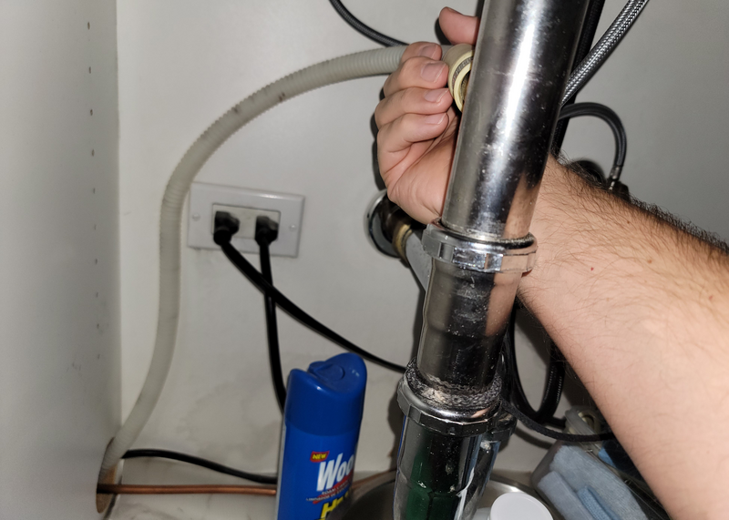 Human hand checks dishwasher hose underneath a kitchen sink with visible plumbing, electrical cords and a spray can of cleaning product, pipe, drainage pipe, metal pipe, white hose, drainage hose, can, blue can, electrical cords, cords, power cords, black cords, under kitchen sink, under sink, kitchen