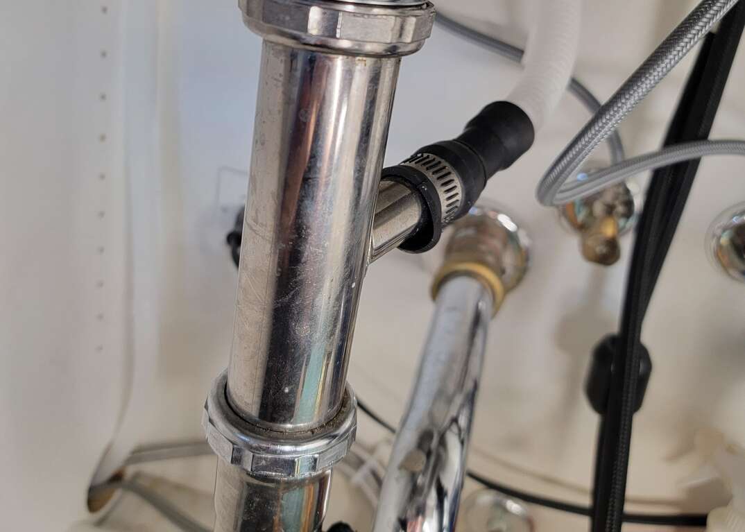 A view under a kitchen sink show a closeup of the air gap hose running from a hole that goes to the dishwasher on the  left side of the frame and runs upward and into the sink drain pipe