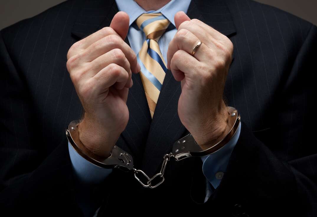 A man in a dark business suit and yellow striped tie with his hands held up in front of the viewer wearing handcuffs around his wrists, business suit, businessman, dark suit, handcuffs, cuffs, arrested, charged, white collar crime