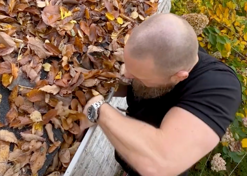 A man standing on a ladder cleans an abundance of colorful fall leaves from the gutters of the roof of a two story house, gutter cleaning, cleaning gutters, gutters, cleaning, ladder, standing on ladder, on ladder, man, male, worker, working, roof, leaves on roof, fall, fall leaves, shrubbery