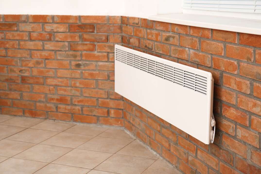 An electric wall heater is affixed to the red brick interior wall of a home with ceramic tile floors, electric wall heater, electric heater, wall heater, red brick interior wall, interior wall, wall, brick, red brick, grout, ceramic tile floor, ceramic tile, tile floor, ceramic floor, heating, HVAC, heater, wall heater