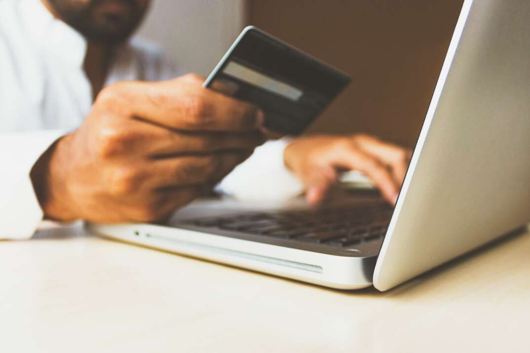 A male wearing a white shirt holds a credit card in one hand while looking at a laptop screen and typing with his other hand, male wearing white shirt, male, white shirt, man, human, human hand, hand, hands, credit card, charge card, credit, finance, financial, finances, laptop computer, laptop, computer, typing, technology, computer monitor, credit score, credit history