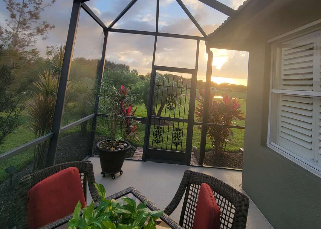 The sunsets in the background from the view out of a screened in porch or lanai in Florida featuring a concrete patio and patio furniture and green plants, lanai, Florida, screened-in porch, porch, patio, patio furniture, sunset, greenery, plants, shutters, window shutters, plantation shutters, concrete, potted plants
