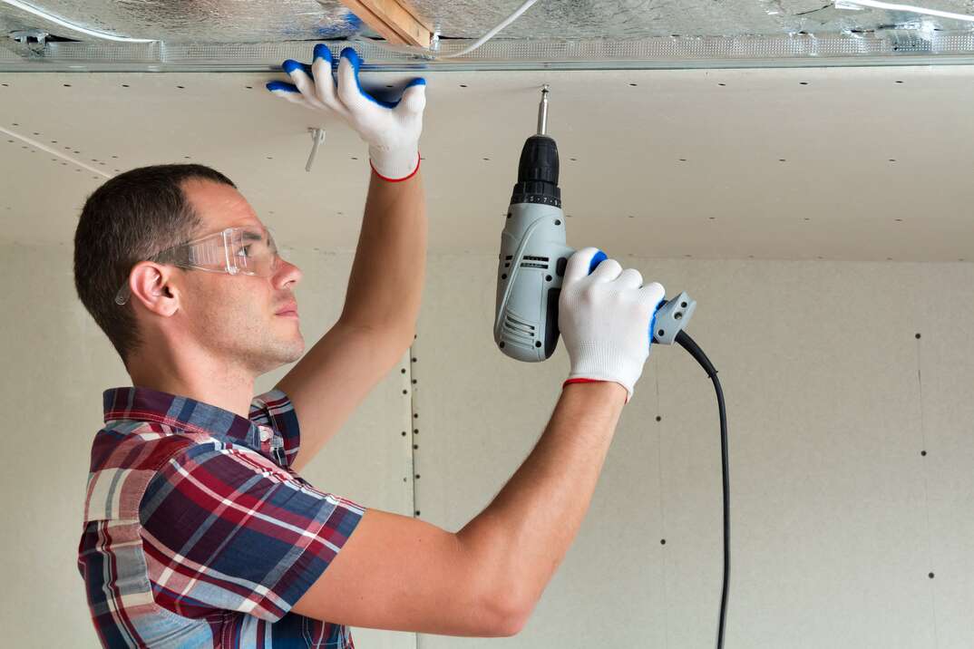 Man in goggles hangs drywall on the ceiling to metal frame using electrical screwdriver