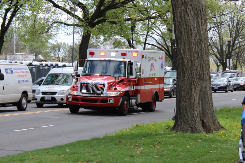 A red and white colored ambulance with its emergency lights on drives on tree lined street past other vehicles, emergency vehicle, vehicle, health care, medical, medical vehicle, medical emergency, emergency, emergency vehicle, street, trees, grass, driving, moving vehicle, emergency lights, flashing lights