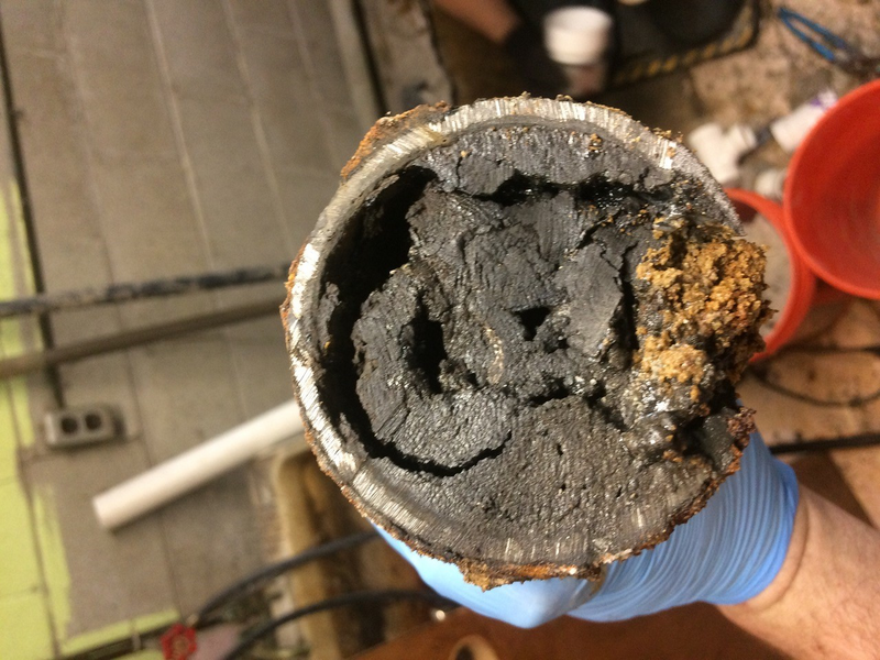 Human hand checks dishwasher hose underneath a kitchen sink with visible plumbing, electrical cords and a spray can of cleaning product, pipe, drainage pipe, metal pipe, white hose, drainage hose, can, blue can, electrical cords, cords, power cords, black cords, under kitchen sink, under sink, kitchen