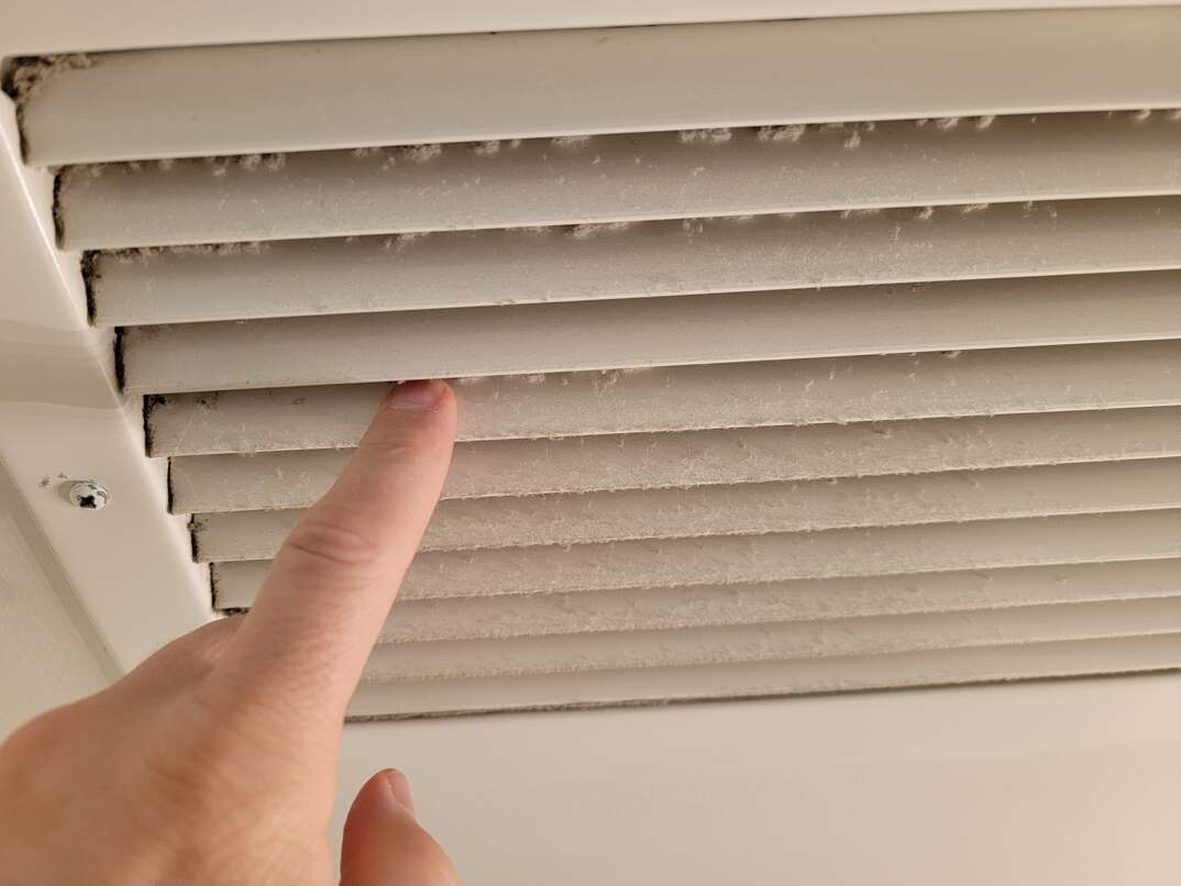 A person's finger points to the dust that has accumulated between the slats of a bathroom fan grate, bathroom fan, bathroom exhaust fan, exhaust fan, fan, bathroom, restroom, dirty, dirty fan, fan grate, grate, filthy, dust, dusty