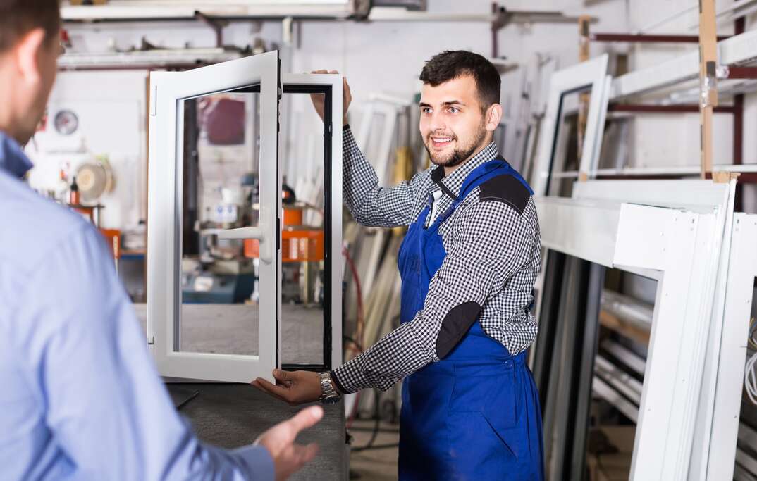 A salesman wearing a blue apron in a window store demonstrates a window for a customer with varied types of other windows surrounding them, windows, window, window shopping, shopping for windows, buying windows, salesman, customer, blue apron, apron, open window, glass