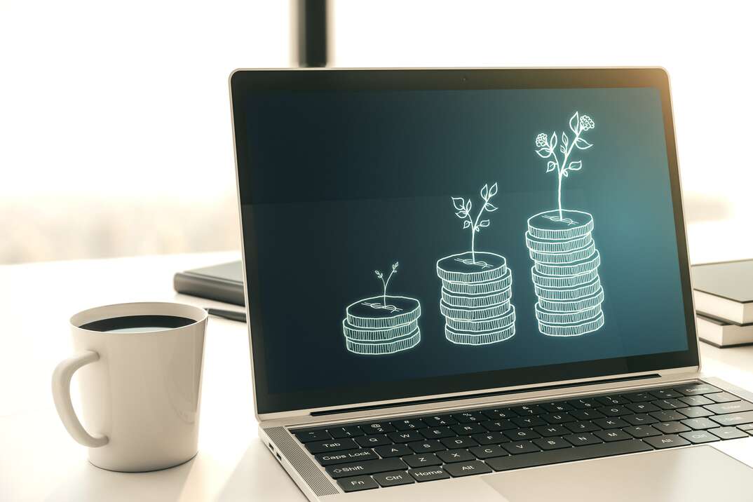 A laptop computer displaying an image of three increasingly taller stacks of coins with increasingly taller trees sprouting out of each sits on a white desk next to a cup of coffee in a brightly lit room, laptop computer, computer screen, computer, laptop, coffee cup, coffee mug, mug, coffee, cup, white desk, desk, stacks of coins, retirement savings, brightly lit room, office