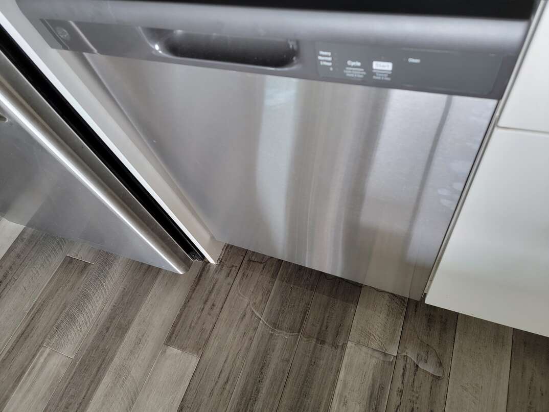 A stainless steel dishwasher leaks onto the gray hardwood flooring in a clean modern kitchen