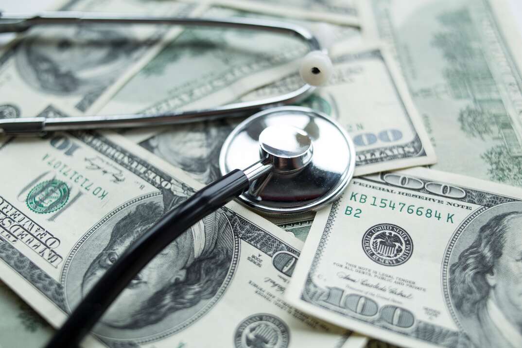 A stethoscope sits on top of a pile of cash in hundred dollar bill denominations, cash, money, hundreds, hundred dollar bills, stethoscope, medical, health coverage, health, insurance, insurance coverage