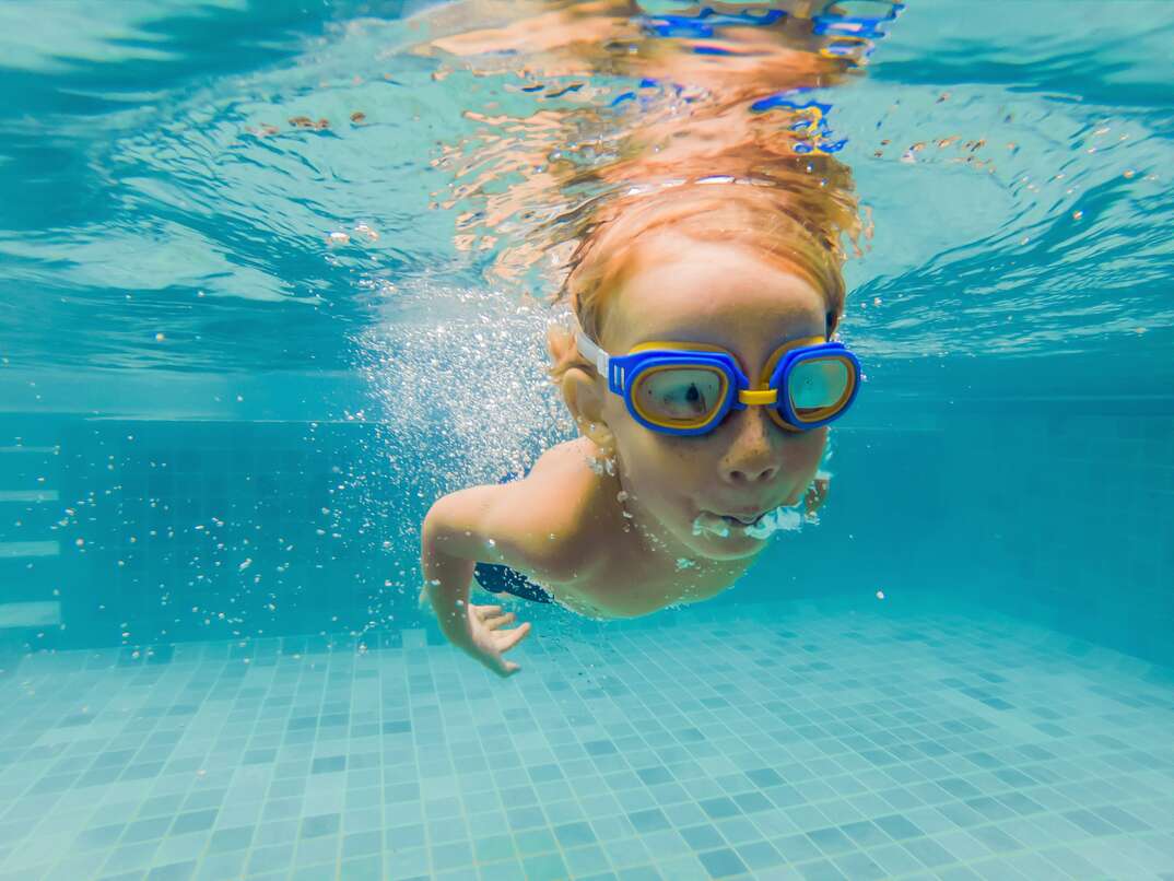 A child boy is swimming underwater in a pool, smiling and holding breath, with swimming glasses.