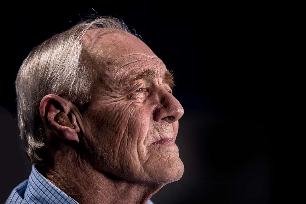 An elderly man with gray hair is displayed in profile against a black background with the hearing aid in his ear visible, hearing aid, hearing, aid, deaf, technology, disabled, disability, handicap, handicapped, gray hair, elderly, elderly man, older man, older, health, health device, device, technology, health coverage, health insurance, health services, ears, ear, black background