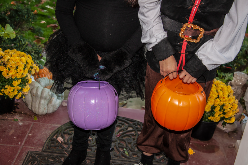 A pair of children wearing Halloween costumes and carrying plastic pumpkin buckets faces the viewer from the vantage of a homeowner handing out candy from their front door, children in Halloween costumers, Halloween costumes, Halloween, costumes, children, kids, plastic pumpkin buckets, pumpkin buckets, pumpkin, buckets, front porch, front stoop, trick-or-treaters, trick-or-treat, Halloween candy