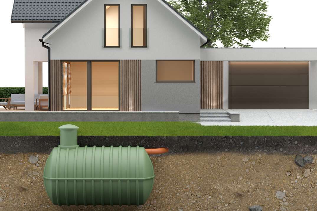 A sideview image shows the profile of both above ground and underground with a green septic tank below the surface of a lawn buried beneath the dirt and grass while the front of a white house with lights on in the windows sits atop it, septic tank, septic sewer, septic, tank, sewer, septic sewer system, sewer system, septic system, plumbing, plumber, dirt, underground, earth, grass, lawn yard, green grass, green lawn, white house, house, home, household, tree, green leaves, lights on, windows