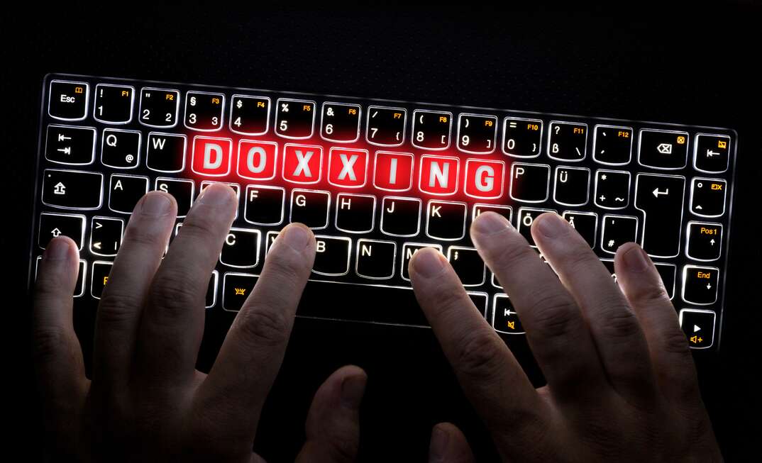 A pair of closeup human hands types on a backlit black computer keyboard with the word doxxing highlighted in red in this photo illustration, doxxing, doxx, dox, keyboard, computer keyboard, computer, laptop, laptop computer, technology, typing, type, fingers, human fingers, hands, human hands, black background