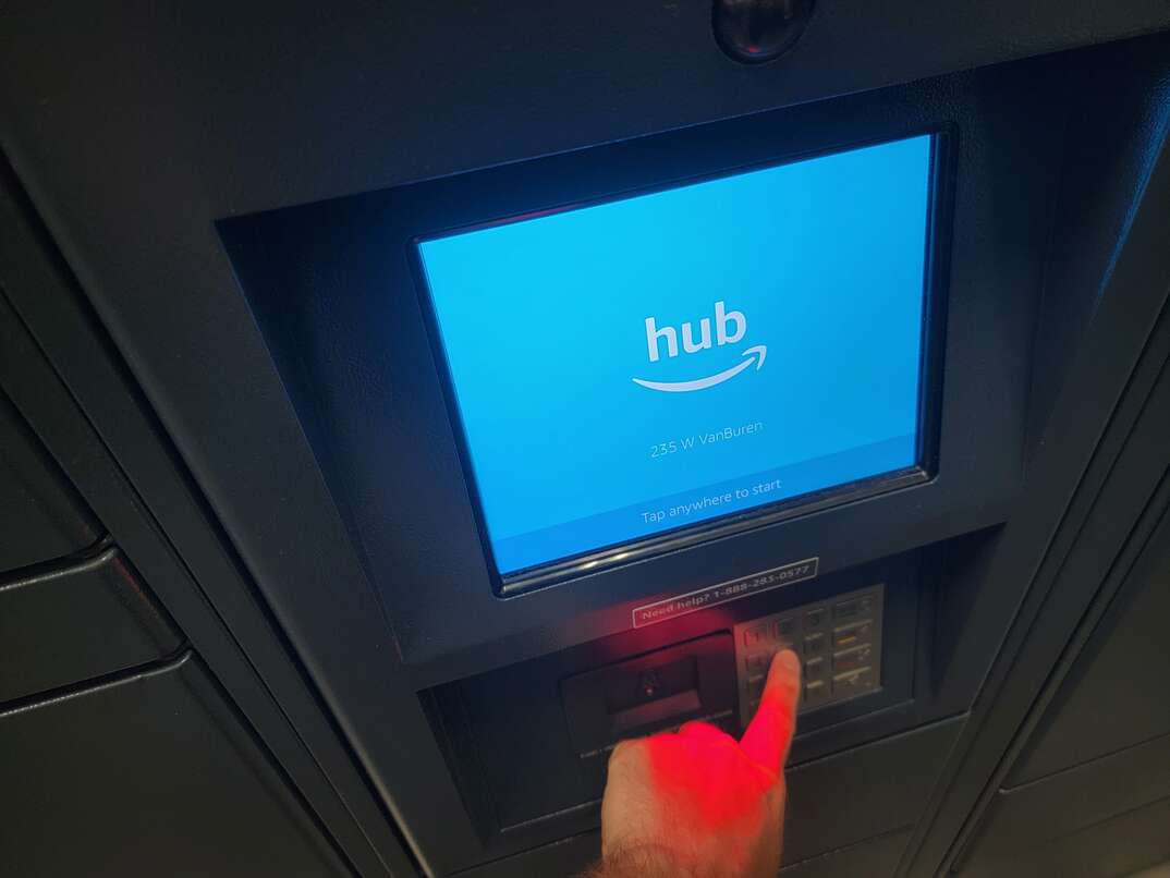 Human hand pressing buttons on the keypad of an Amazon Hub package system with a blue screen bearing the Hum log
