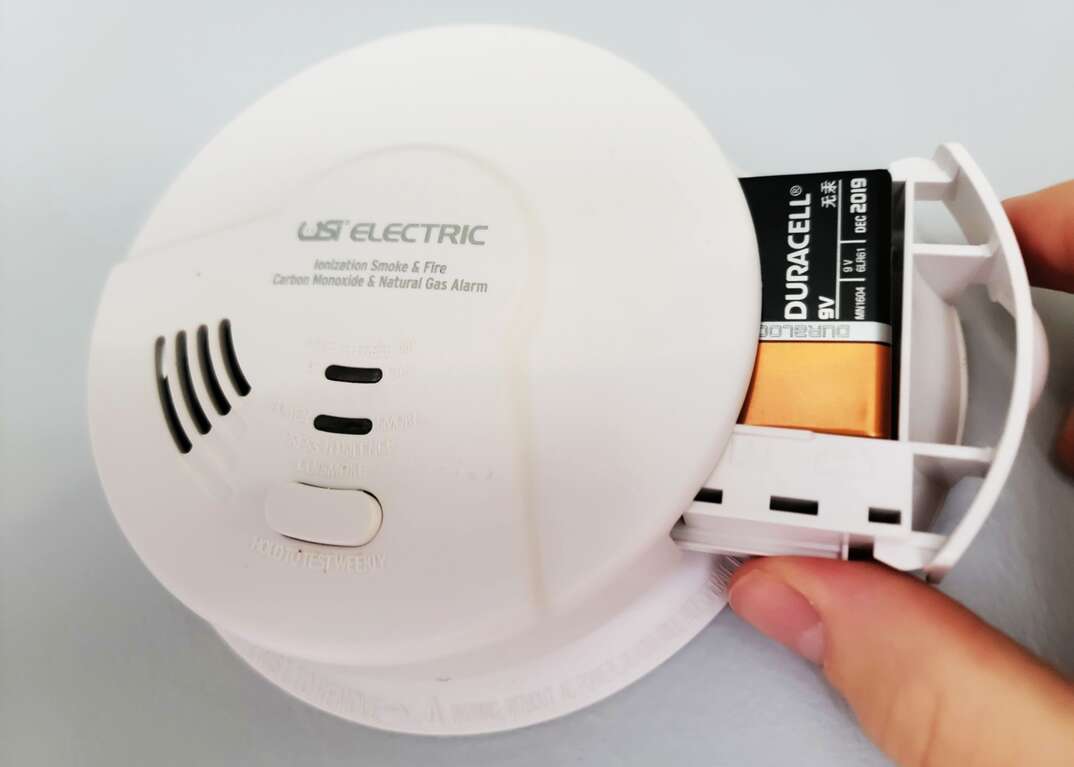 A human hand replaces the 9-volt battery in a home smoke alarm and carbon monoxide detector